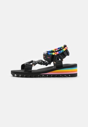 Sandalia de cuña negra con tiras ajustables, pulsera de tobillo con cuentas de colores y suela con plataforma a rayas de arcoíris.