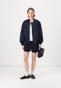 Ναυτικό bomber μπουφάν με φερμουάρ και ριπ μανσέτες, συνδυασμένο με αντίστοιχες βερμούδες. Λευκό πουκάμισο από κάτω, κρατώντας μια μαύρη τσάντα, με σκούρα παπούτσια.