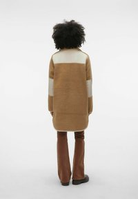 Cappotto in pile marrone con colletto alto, caratterizzato da un pannello crema sulle spalle e maniche lunghe. Indossato sopra pantaloni marroni a zampa d'elefante e stivali.