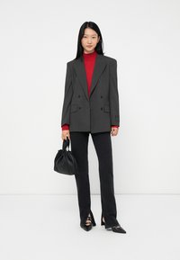 Frau in einem grauen Nadelstreifenblazer, rotem Rollkragenpullover, schwarzen Hosen mit Schlitzsäumen, schwarzen spitzen Schuhen und einer schwarzen Lederhandtasche.
