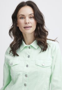 Chaqueta de mezclilla verde menta con cuello, botones a presión plateados y bolsillos dobles en el pecho. Presenta un diseño ajustado y una tela texturizada.