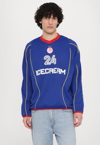 Blauw shirt met rode accenten, met het nummer "24" en "ICECREAM" in het wit. Voorzien van gele stiksels en een ronde halslijn.