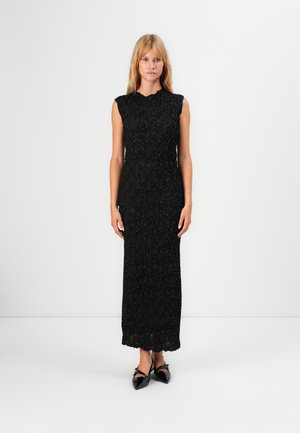 Donna in piedi con indosso un maxi abito senza maniche nero a trama e scarpe con tacco a punta nere, contro uno sfondo bianco uniforme.