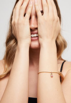 Jeune femme aux cheveux ondulés se couvrant le visage avec les mains, souriante, portant un bracelet de perles dorées avec un charme rouge au poignet gauche.