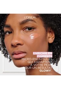 Fond de teint crémeux avec une formule radieuse, appliqué sur la joue ; le modèle a la peau brun moyen et des boucles naturelles. Texte en français inclus.