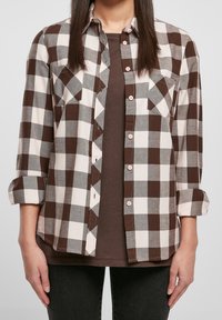 Camicia a quadri marroni e crema con due tasche frontali, design con bottoni e maniche arrotolate. Indossata sopra una maglietta marrone, abbinata a jeans scuri.