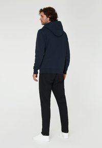 Sudadera navy con un bolsillo frontal y capucha con cordón, combinada con pantalones negros y zapatillas blancas. Presenta un tejido texturizado y un diseño minimalista.