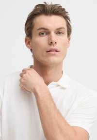 Polo blanc avec col, tissu texturé, manches courtes. La main du modèle est posée sur l'épaule, mettant en valeur un fond neutre.