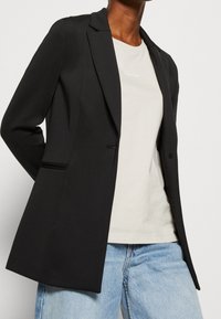 Czarna marynarka z gładkiej tkaniny, z klapą z wycięciem, pojedynczym zapięciem na guzik oraz bocznymi kieszeniami, zestawiona z jasnym t-shirtem i dżinsami.