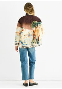 Quilted jacket featuring a brown gradient yoke, detailed with colorful landscape artwork and a text border along the bottom. Worn with blue jeans.