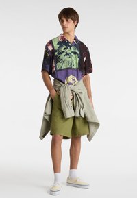 Kortærmet skjorte med multikolorede abstrakte designs i grønne og lilla nuancer. Sammen med olivengrønne shorts og en lys grå jakke bundet om livet.