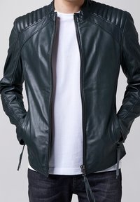 Giacca in pelle verde scuro con accenti a coste sulle spalle, chiusura con zip e tasche laterali. Indossata sopra una camicia bianca e jeans scuri.