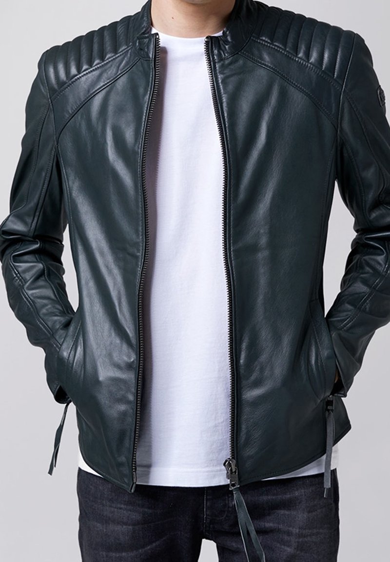 Giacca in pelle verde scuro con accenti a coste sulle spalle, chiusura con zip e tasche laterali. Indossata sopra una camicia bianca e jeans scuri.