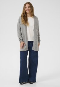 Hellgrauer Cardigan aus weichem Material, getragen über einem weißen Hemd, kombiniert mit weit geschnittenen, dunkelblauen Jeans und schwarzen, genoppten Schuhen.