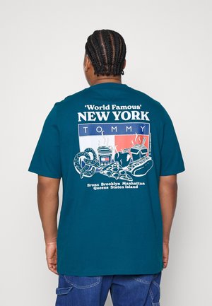 Person med fletninger iført en teal T-shirt, der viser rygdesignet af New York-mad og bydele navne i hvidt, rødt og blåt.