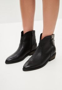 Botoși din piele neagră până la gleznă, cu vârf ascuțit, fermoar pe lateral, accente texturate și toc mic, pătrat. Design cu model pe partea superioară.