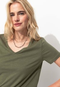 T-shirt vert olive en V fabriqué dans un tissu doux avec des manches courtes. Il est agrémenté d'un collier en perles argentées superposées. Design simple avec une légère texture.
