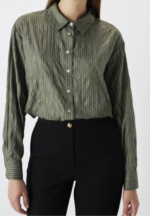 Kvinde iført en langærmet, tekstureret olivengrøn skjorte med knapper, stoppet ned i højtaljede sorte bukser med en guldknap.