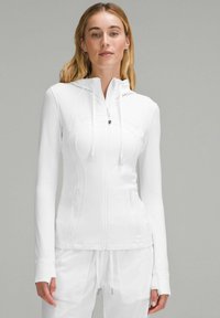 Haut à zip blanc avec une coupe ajustée, doté de manches longues, d'une capuche et de deux poches zippées. Texture lisse et détails de couture sans couture.