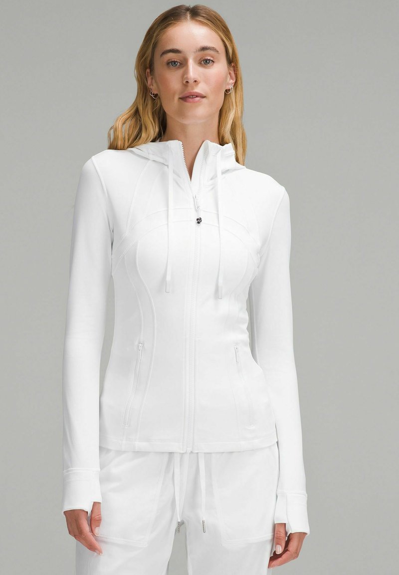 Haut à zip blanc avec une coupe ajustée, doté de manches longues, d'une capuche et de deux poches zippées. Texture lisse et détails de couture sans couture.