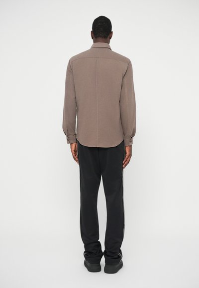 Brun langermet skjorte med krage, med rett kant og knyttede ermer, iført svarte joggebukser og mørke sko.