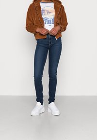 Hnědá kordová bunda s kapucí přes bílým grafickým tričkem. Úzké tmavě modré džíny a bílé platformové tenisky doplňují celý outfit.