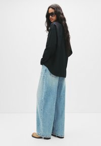 Modella che indossa una camicia nera oversize con maniche lunghe, abbinata a jeans a gamba larga di colore blu chiaro con macchie di vernice. Occhiali da sole neri e scarpe beige.