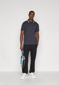 Σκούρο πόλο μπλουζάκι με λευκό γιακά, σε συνδυασμό με μαύρο υφασμάτινο παντελόνι. Λευκά αθλητικά παπούτσια και ρακέτα τένις. Απλός και λειτουργικός σχεδιασμός.