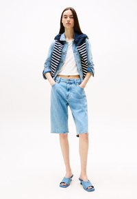 Veste en denim avec un pull rayé bleu marine et blanc drapé sur les épaules, haut blanc, shorts en denim d'un lavage clair et chaussures bleu ciel à enfiler.