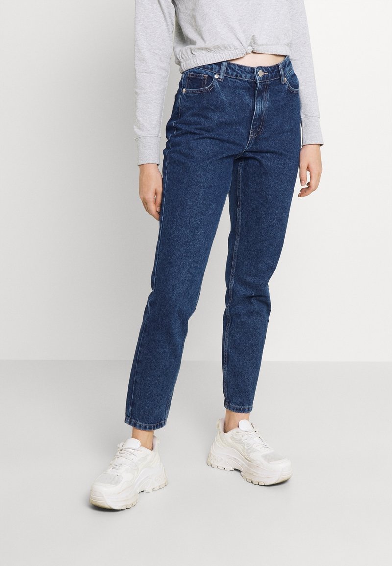 Jeans en denim bleu foncé avec une taille haute, un design à jambe droite et des ourlets retroussés, associés à des chaussures de sport blanches et un haut gris clair.