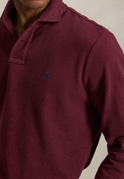 Homme portant un polo à manches longues rouge foncé avec un petit logo brodé bleu foncé sur la poitrine.