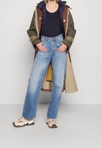 Långa ljusblå jeans med rak passform, kombinerade med en mörk tanktopp och en lager-på-lager olivgrön och brun jacka. Bär beiga träningsskor.