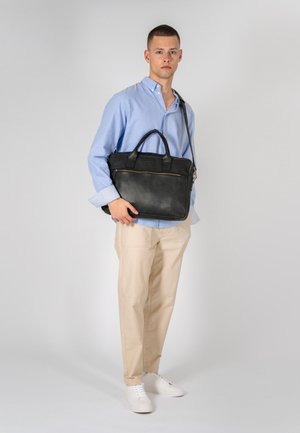 Maletín de cuero negro con asas superiores y correa de hombro ajustable, que cuenta con un bolsillo frontal con cremallera y una textura suave. El modelo lleva una camisa y pantalones claros.