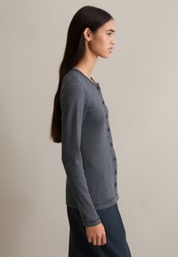 Szary kardigan na guziki z długimi rękawami, o prążkowanej fakturze, z okrągłym dekoltem i kontrastowymi przeszyciami. Noszony na ciemnych spodniach.