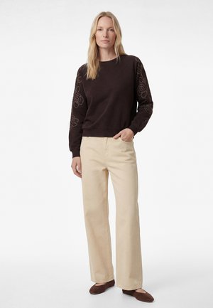 Kobieta ubrana w ciemną bluzę z długim rękawem z kwiatowym haftem na rękawach, beżowe spodnie o szerokich nogawkach oraz brązowe baleriny, stoi z jedną ręką w kieszeni.