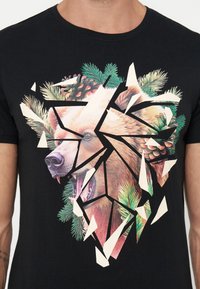 Tête d'ours rugissante avec des pommes de pin et des branches persistantes, fragmentée en éclats géométriques sur le devant d'un t-shirt noir.