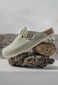 Zoccolo verde in camoscio con fibbia dorata, caratterizzato da una punta arrotondata e una soletta testurizzata, adagiato su rocce testurizzate, che mette in risalto un design minimalista.
