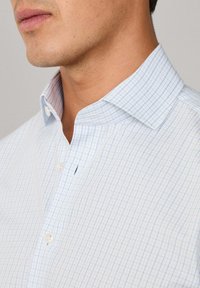 Muškarac nosi svijetloplavu i bijelu košulju s kariranim uzorkom, s otvorenim ovratnikom, koji otkriva dio njegove čeljusti i vrata.