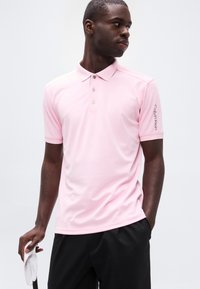 Άνδρας με ροζ πόλο πουκάμισο Calvin Klein και μαύρο παντελόνι κρατά λευκό γάντι γκολφ, κοιτάζοντας προς το πλάι μπροστά σε απλό φόντο.