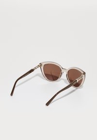 Ochelari de soare tip cat-eye cu rame de culoare roz deschis, lentile nuanțate de maro și detalii aurii complicate pe brate. Brațele sunt din acetat maro.