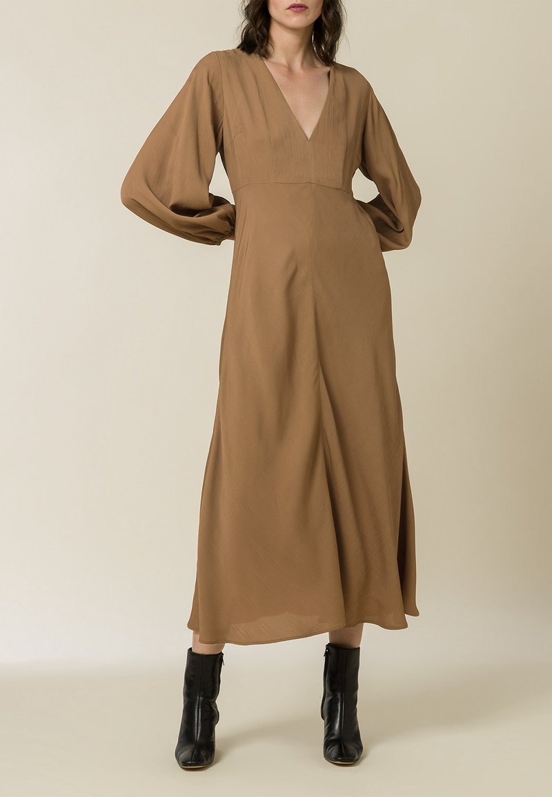 Femme portant une longue robe marron avec un col en V et des manches longues bouffantes, associée à des bottines noires, debout les mains sur les hanches.