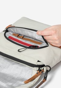 Hand holding open beige bag’s zippered pocket showing gray grid-lined interior, small red zippered compartment, and brown leather straps.