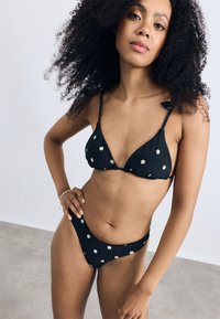 Svart bikini-set med en triangulär topp och högskurna bottoms, båda prydda med vit blomstermönstring. Tillverkad av ett mjukt tyg.