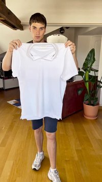Camisa polo blanca sostenida por una persona, con un cuello clásico y mangas cortas. Textura suave, diseño casual, fondo con plantas de interior.