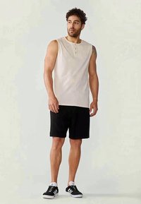 Beige sleeveless henley shirt with three buttons, paired with black shorts and black sneakers featuring white accents. Lightweight and casual fit.