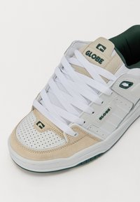 Men's sneaker with white leather and beige suede upper, green accents, perforated toe, and white laces. Includes a rubber outsole for grip.