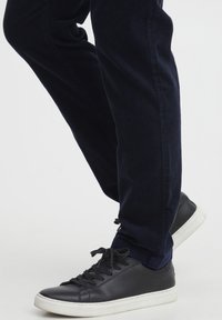 Sneaker in pelle nera con suola in gomma bianca, abbinati a pantaloni slim-fit blu scuro. Le sneaker hanno una texture liscia e dettagli con lacci.
