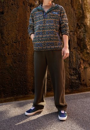 Person trägt gemusterten Fleecepullover mit Columbia-Logo, lockere braune Fleecehose und marineblaue Sneaker und steht auf dem Gehweg neben einer strukturierten Wand.