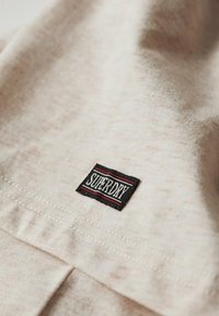 Beige cotton fabric with a textured surface, featuring a black patch with "SUPERDRY" in white letters and red accents. Seam detail visible.