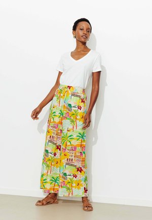 Femme portant un T-shirt blanc à col en V, un pantalon large coloré à imprimé tropical, des boucles d'oreilles en forme de créoles dorées, et des sandales marron cloutées, debout contre un mur blanc.